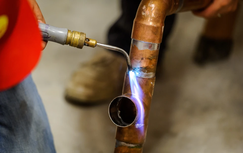 Un plombier en veste rouge utilise un chalumeau à gaz pour souder une canalisation en cuivre dans un environnement intérieur