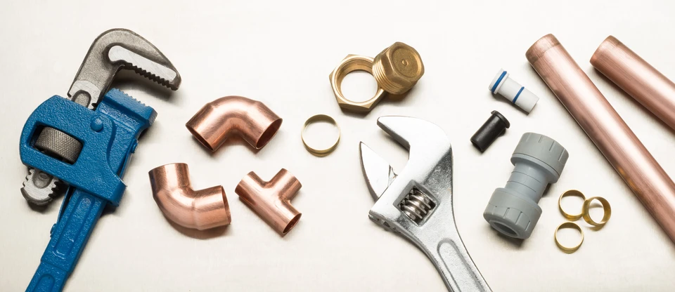 Outils de plomberie disposés sur surface blanche : clé à molette bleue, clé anglaise chromée, raccords en cuivre coudés et en T, raccords laiton filetés, manchons PVC gris, tube cuivre, bagues d'étanchéité dorées