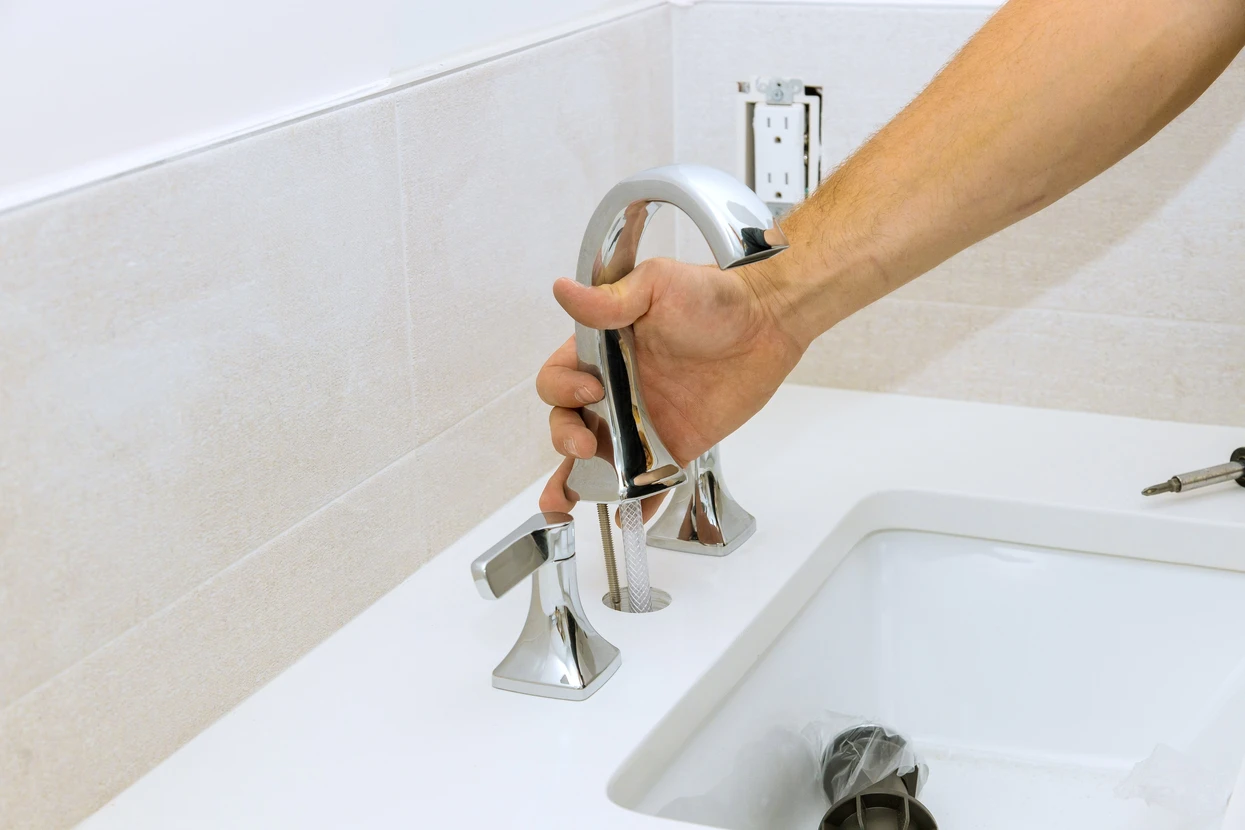 Un plombier en combinaison bleue et gants de protection installe ou répare un robinet de baignoire chromé dans une salle de bain moderne aux murs carrelés blancs