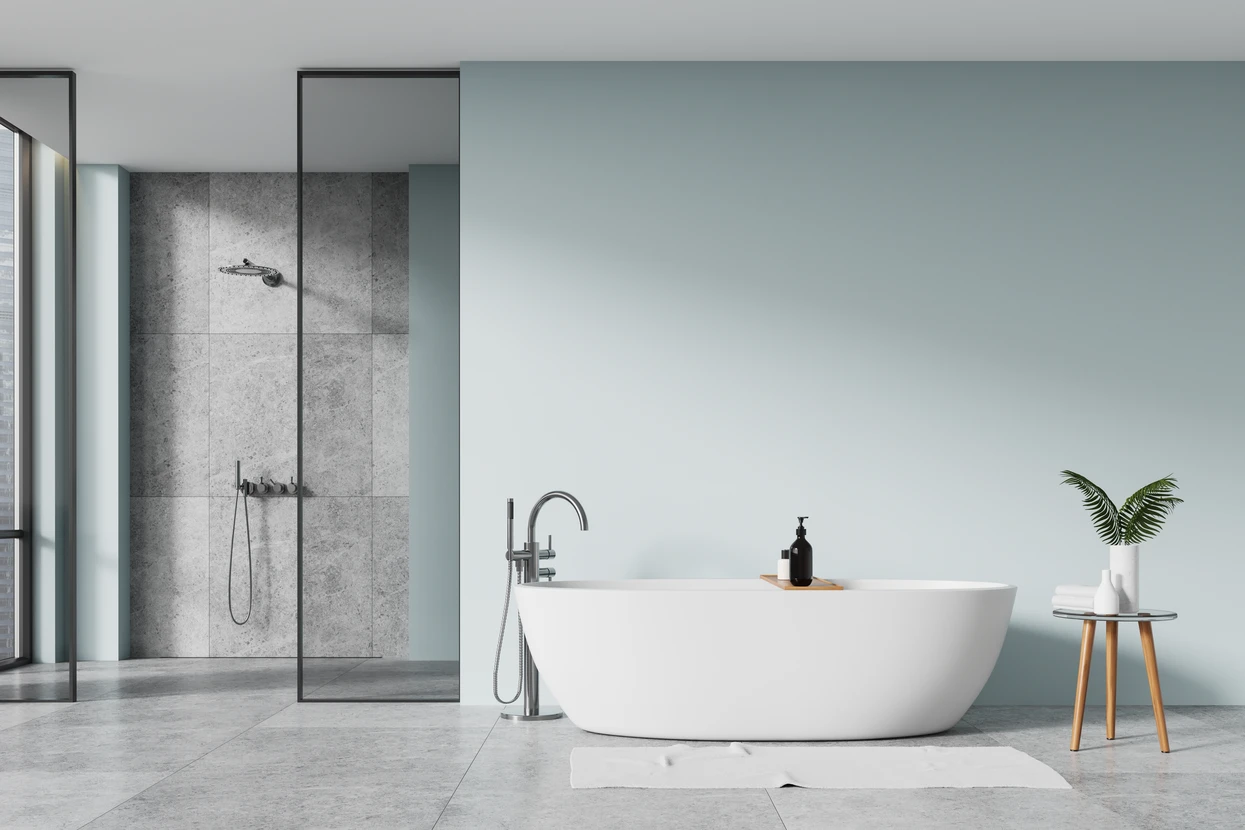 Un plombier en combinaison bleue et gants de protection installe ou répare un robinet de baignoire chromé dans une salle de bain moderne aux murs carrelés blancs