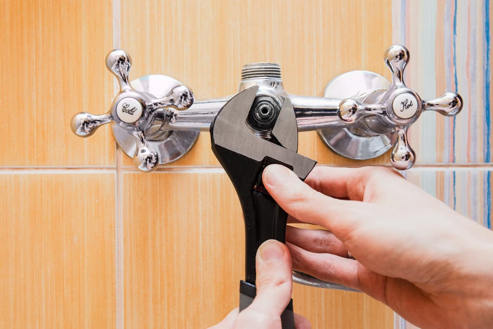Un plombier en combinaison de travail grise et bleue installe ou répare une cuvette de toilettes suspendue dans une salle de bains moderne aux murs carrelés blancs