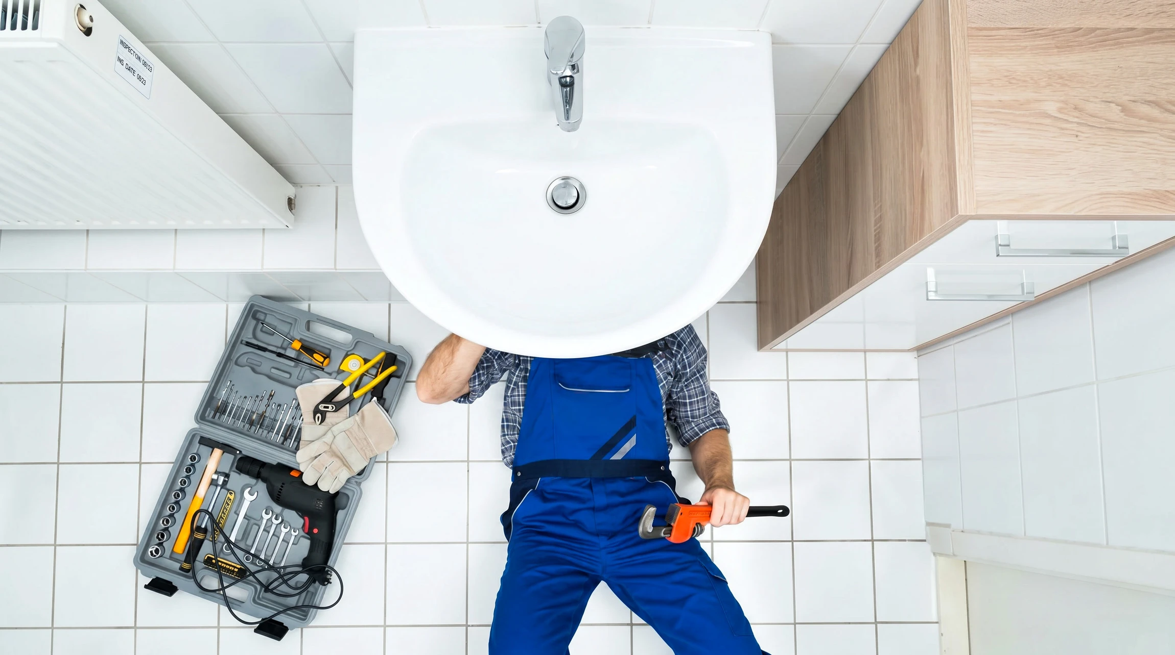 Plombier en combinaison bleue intervenant sous un évier de cuisine, ajustant une canalisation avec une clé à molette